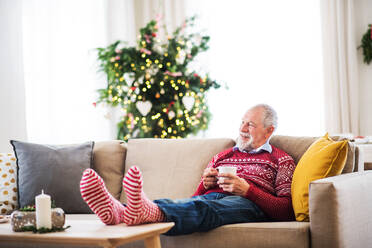 Ein älterer Mann mit den Füßen auf einem Couchtisch, der zu Hause auf dem Sofa sitzt und eine Tasse Kaffee in der Hand hält, zur Weihnachtszeit. - HPIF29319