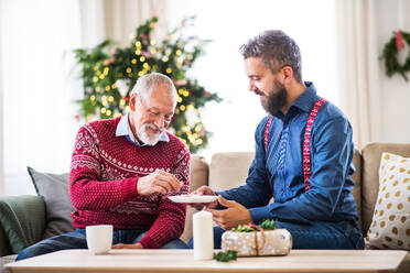 A senior father and adult son sitting on a sofa at home at Christmas time, eating biscuits and talking. - HPIF29321