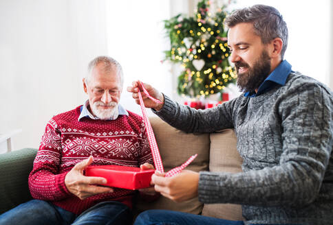 Ein älterer Vater und sein erwachsener Sohn sitzen zur Weihnachtszeit zu Hause auf dem Sofa und packen ein Geschenk ein. - HPIF29332