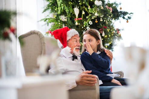 Ein kleines Mädchen und ihr Großvater mit Weihnachtsmannmütze sitzen zur Weihnachtszeit auf einem Sessel und erzählen ein Geheimnis. - HPIF29365