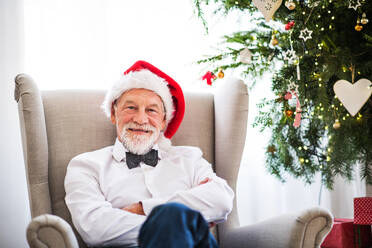 A senior man with a Santa hat sitting on an armchair at home at Christmas time. - HPIF29370