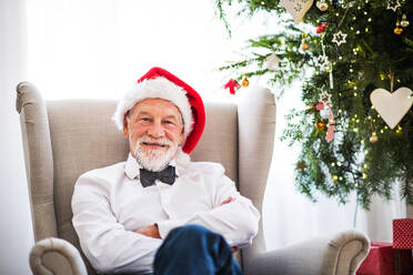 Ein älterer Mann mit einer Weihnachtsmannmütze sitzt zur Weihnachtszeit zu Hause auf einem Sessel. - HPIF29370