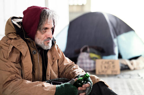 A homeless beggar man sitting outdoors in city asking for money donation, holding a bottle of alcohol. Copy space. - HPIF29431