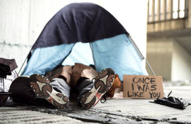 Beine und Füße eines obdachlosen Bettlers, der draußen in der Stadt auf dem Boden liegt, in einem Zelt schläft und um eine Geldspende bittet. - HPIF29436