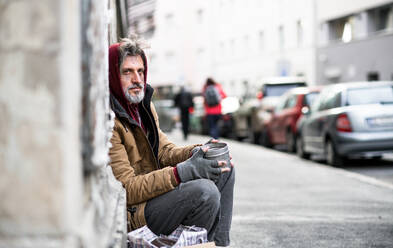 Seitenansicht eines obdachlosen Bettlers, der vor einem Holztor in der Stadt sitzt und um eine Geldspende bittet. - HPIF29478