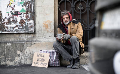 Ein obdachloser Bettler sitzt vor einem Holztor in der Stadt und bittet um eine Geldspende. - HPIF29480