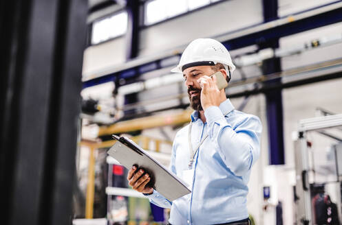 Porträt eines reifen Industrieingenieurs mit Klemmbrett und Smartphone in einer Fabrik, der einen Anruf tätigt. Kopierraum. - HPIF29497