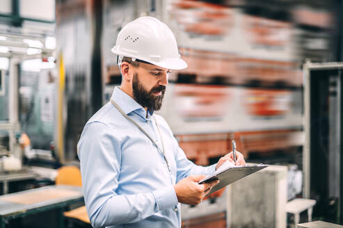 Porträt eines reifen Industrieingenieurs mit Klemmbrett in einer Fabrik, schreibend, Raum kopieren. - HPIF29500