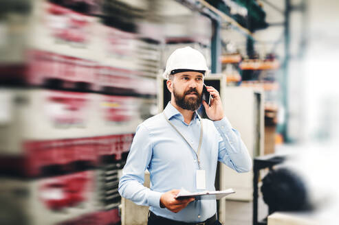 Porträt eines reifen Industrieingenieurs mit Klemmbrett und Smartphone in einer Fabrik, der einen Anruf tätigt. Kopierraum. - HPIF29501