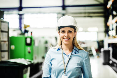 A portrait of a young industrial woman engineer with white helmet standing in a factory. - HPIF29503