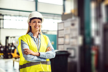 Das Porträt einer jungen Ingenieurin, die mit verschränkten Armen in einer Fabrik steht. - HPIF29504