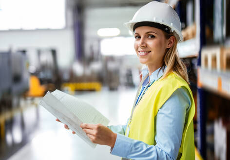 Das Porträt einer jungen Ingenieurin, die in einer Fabrik steht und Baupläne in der Hand hält. - HPIF29506