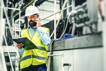 Porträt eines reifen Industrieingenieurs mit Klemmbrett und Smartphone in einer Fabrik, der einen Anruf tätigt. Kopierraum. - HPIF29544