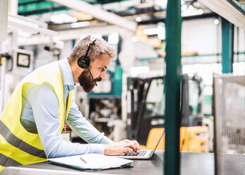 Porträt eines reifen Industrieingenieurs mit Headset und Laptop in einer Fabrik, bei der Arbeit. - HPIF29551