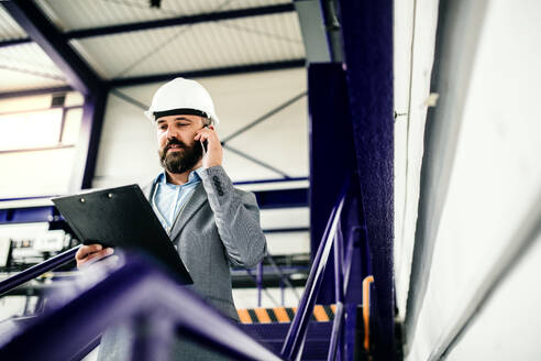 Porträt eines reifen Industrieingenieurs mit Klemmbrett und Smartphone in einer Fabrik, der einen Anruf tätigt. Kopierraum. - HPIF29557