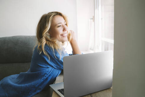 Glückliche Frau sitzt mit Laptop am Fenster zu Hause - NDEF00623