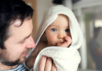 Vater mit einem glücklichen Kleinkind, das in ein Handtuch eingewickelt ist, in einem Badezimmer zu Hause. Vaterschaftsurlaub. Nahaufnahme. - HPIF29636