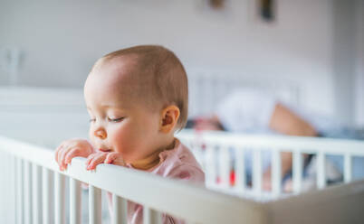 A happy toddler girl standing in a cot in the bedroom at home. Unrecognizable father in the background. Close up. - HPIF29664