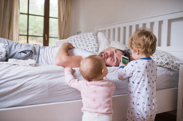 Zwei Kleinkinder, die mit dem Smartphone spielen, und ihr Vater, der auf dem Bett im Schlafzimmer liegt. Vaterschaftsurlaub. - HPIF29677