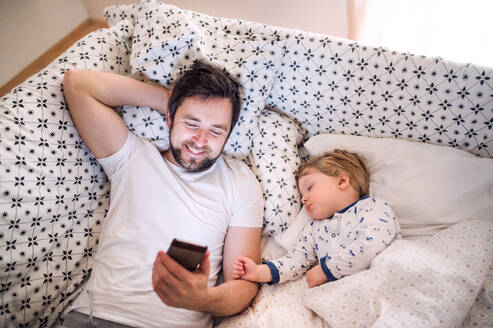 Vater mit Smartphone und einem schlafenden Kleinkind im Bett zu Hause. Vaterschaftsurlaub. - HPIF29684