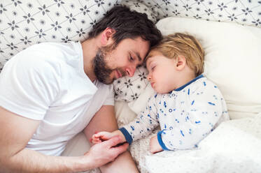 Father holding hand of a sleeping toddler boy lying in bed at home. Paternity leave. - HPIF29686