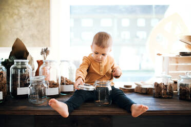 Ein glücklicher barfuß laufender kleiner Junge sitzt auf einem Tisch in einem Zero-Waste-Laden und hält ein Glas in der Hand. - HPIF29715