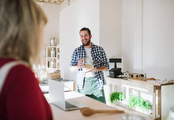 Junger männlicher Verkäufer mit Laptop, der eine nicht erkennbare Frau in einem Zero Waste Shop bedient. - HPIF29724