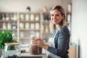 A young female shop assistant in a zero waste shop standing at the counter, holding a jar with groceries. - HPIF29757