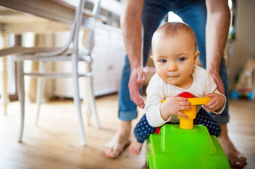 Unrecognizable father with a baby girl sitting on a toy car at home. Paternity leave. - HPIF29790