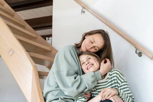 Zärtliche Mutter, die ihre Tochter umarmt, während sie zu Hause auf den Stufen sitzt - NDEF00658