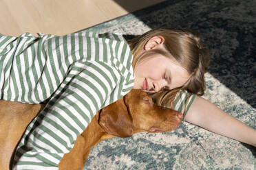 Mädchen liegt mit Hund auf Teppich zu Hause an einem sonnigen Tag - NDEF00659