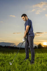 Junger Mann bereitet sich auf einen Golfschlag auf Gras in der Abenddämmerung vor - STSF03723