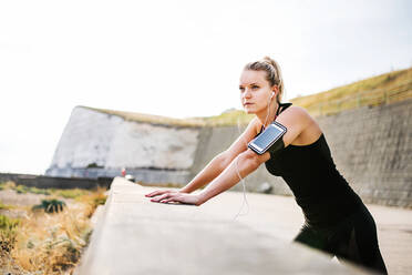 Young sporty woman runner with earphones and smartphone in armband standing outside on the beach in nature, listening to music and resting. - HPIF29861