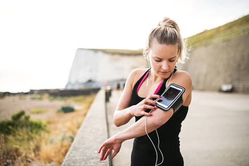 Junge sportliche Läuferin mit Kopfhörern, die draußen am Strand in der Natur steht und ihr Smartphone in der Armbinde benutzt. - HPIF29862