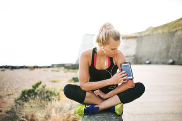 Junge sportliche Frau Läuferin mit Kopfhörer sitzen draußen am Strand in der Natur, mit Smartphone in Armbinde. - HPIF29865