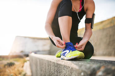 Unrecognizable young sporty woman runner with earphones and smartphone in an armband outside by the beach, tying shoelaces. - HPIF29866