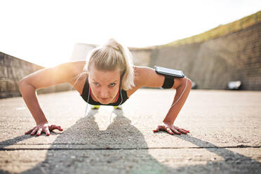 Young sporty woman runner with earphones and smartphone in armband doing push-ups outside in nature, listening to music. - HPIF29868