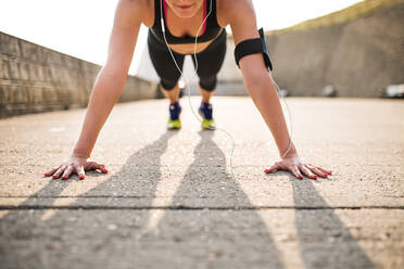 Unrecognizable young sporty woman runner with earphones and smartphone in armband doing push-ups outside in nature, listening to music. - HPIF29869