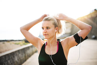 Junge sportliche Läuferin mit Kopfhörern und Smartphone in der Armbinde steht draußen am Strand in der Natur, hört Musik und ruht sich aus. - HPIF29871