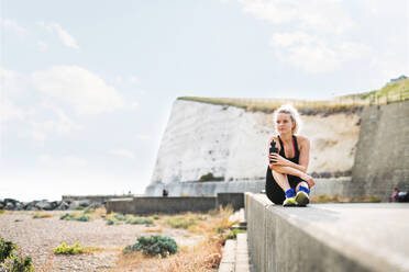 Young sporty woman runner with water bottle sitting outside on the beach in nature, resting. Copy space. - HPIF29872
