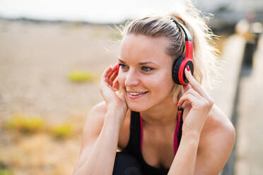 A close-up of young woman runner with black and red headphones resting outside by the sea, listening to music. - HPIF29873