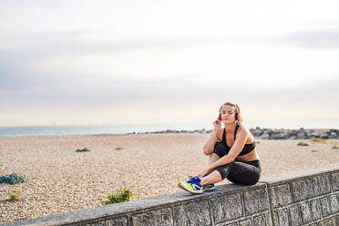 A young woman runner with black and red headphones resting outside by the sea, listening to music. - HPIF29874