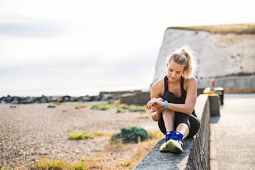 Junge sportliche Läuferin, die sich draußen in der Natur ausruht, auf einer Mauer sitzt und ihre Smartwatch einstellt. - HPIF29875