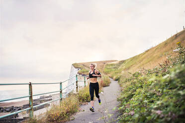 Young sporty woman runner with earphones running by the sea outside in nature, listening to music. - HPIF29876