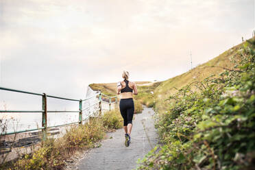A rear view of young sporty woman runner with smartphone in an armband running by sea in nature. - HPIF29877