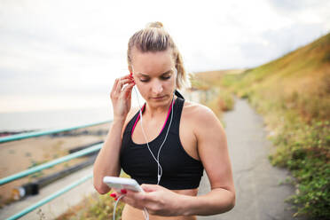 Junge sportliche Läuferin mit Kopfhörern und Smartphone steht draußen am Strand in der Natur, hört Musik und ruht sich aus. - HPIF29880