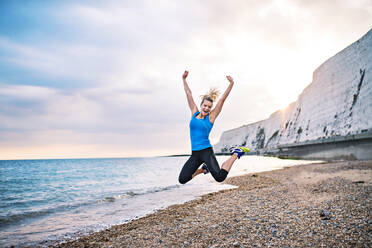 Junge sportliche Läuferin in blauer Sportkleidung, die draußen am Strand in der Natur springt. copy space. - HPIF29893