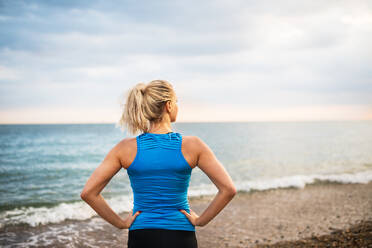 Junge sportliche Läuferin in blauer Sportkleidung steht draußen am Strand in der Natur und ruht sich aus, Rückansicht. - HPIF29898