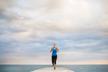 A young sporty woman running outside on a pier, by the ocean. Copy space. - HPIF29909