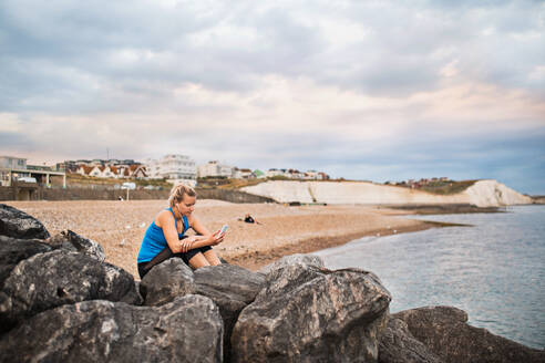 Junge sportliche Läuferin mit Kopfhörern und Smartphone sitzt draußen am Strand in der Natur, hört Musik und ruht sich aus. - HPIF29910